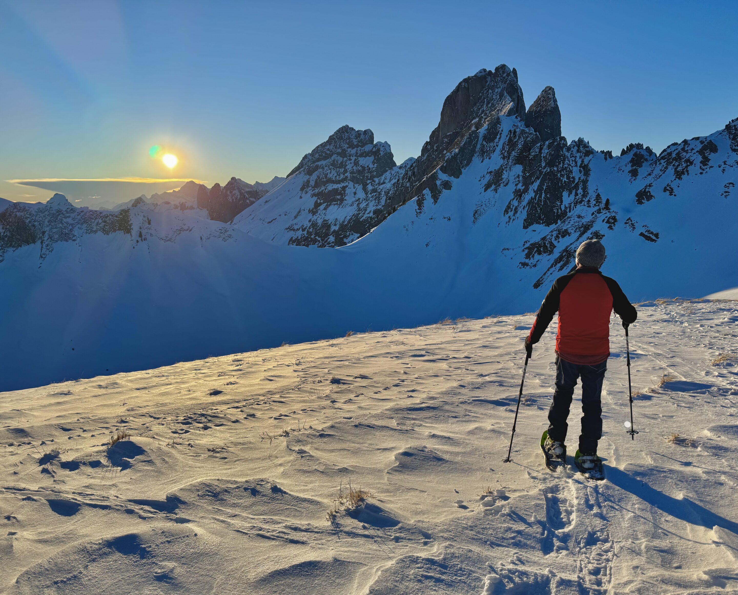 Person beim Schneewandern bei Sonnenuntergang in den verschneiten Bergen, mit beeindruckendem Bergpanorama im Hintergrund.