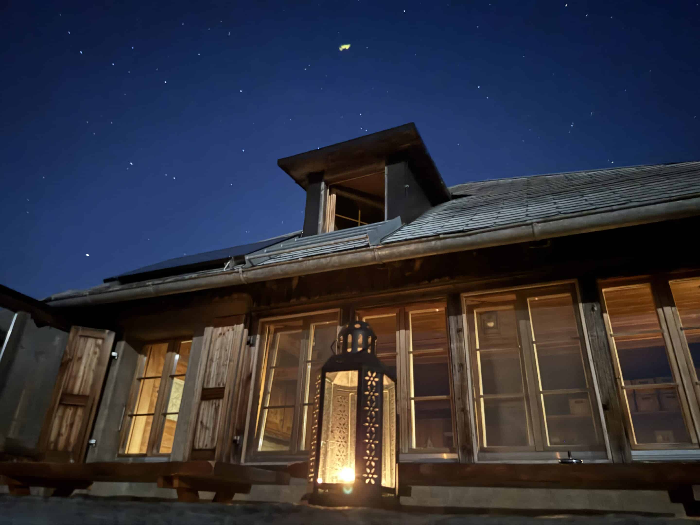 Rustikales Holzhaus bei Nacht mit beleuchteter Laterne und sternenklarem Himmel.