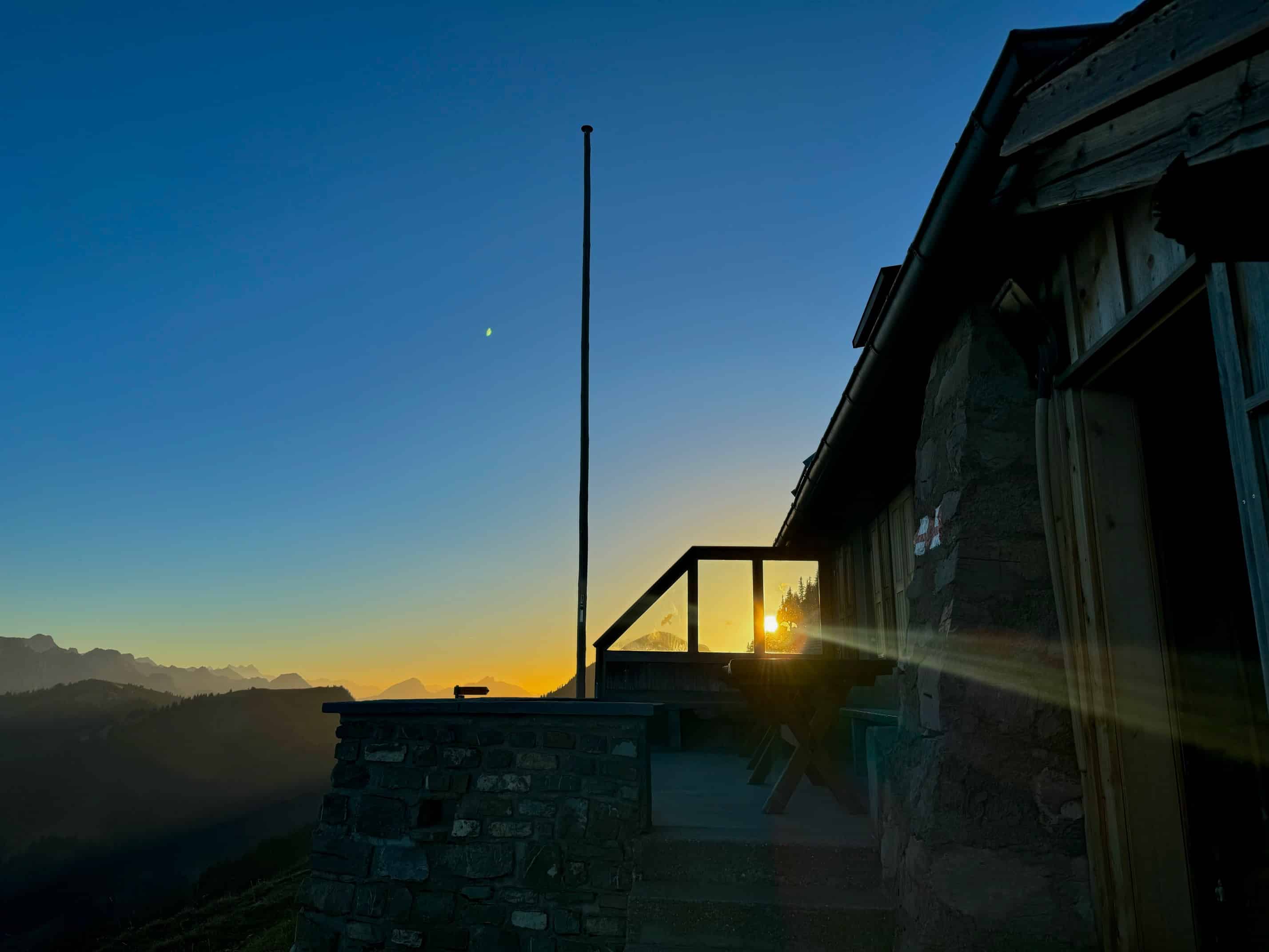 Berghütte im Sonnenuntergang mit klarer Himmel und Bergsilhouette im Hintergrund, ruhige Abendstimmung.