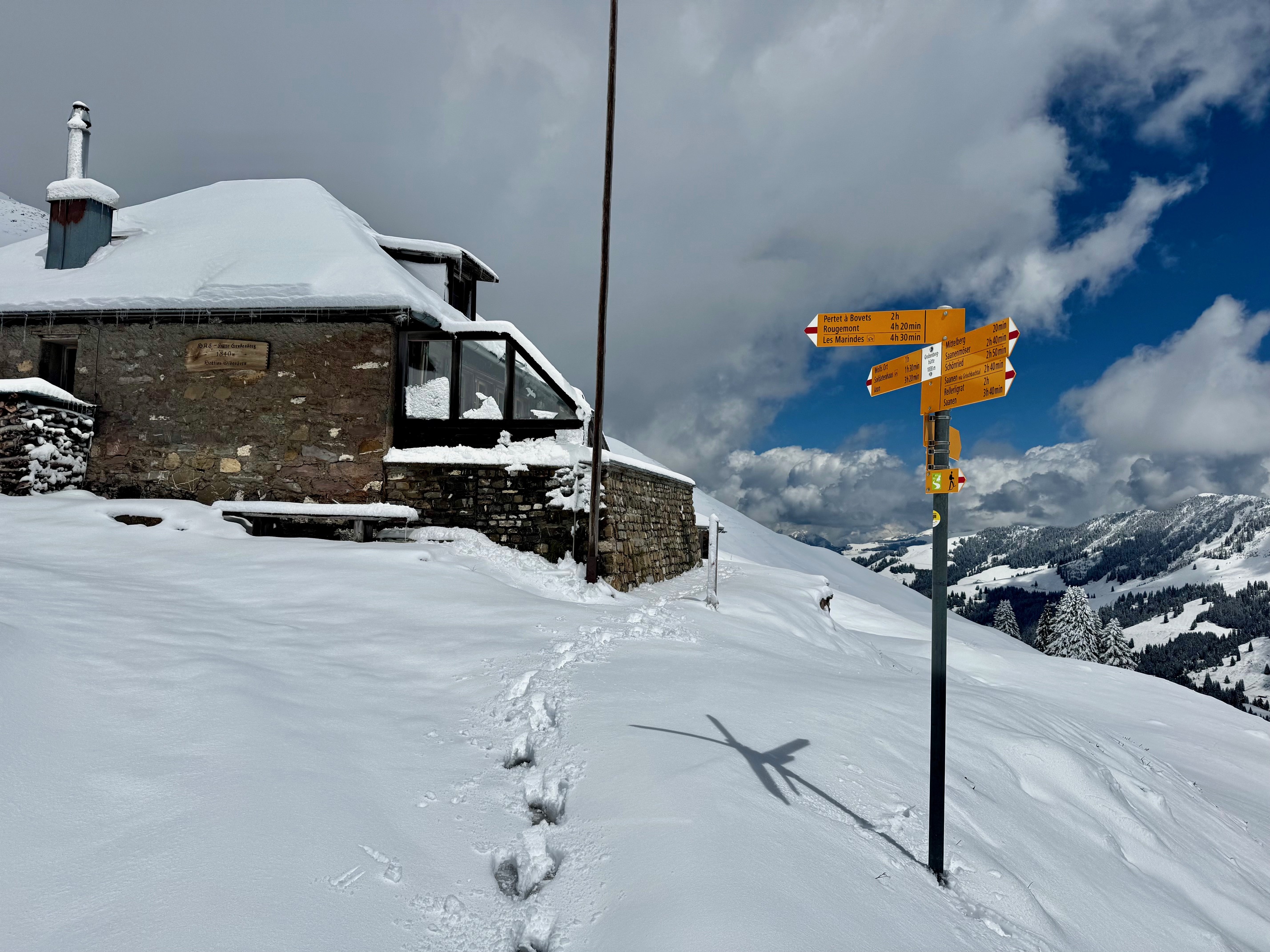 IMG_0522 Schneebedeckte Berghütte mit Wegweiser, umgeben von winterlicher Alpenlandschaft und bewölktem Himmel.