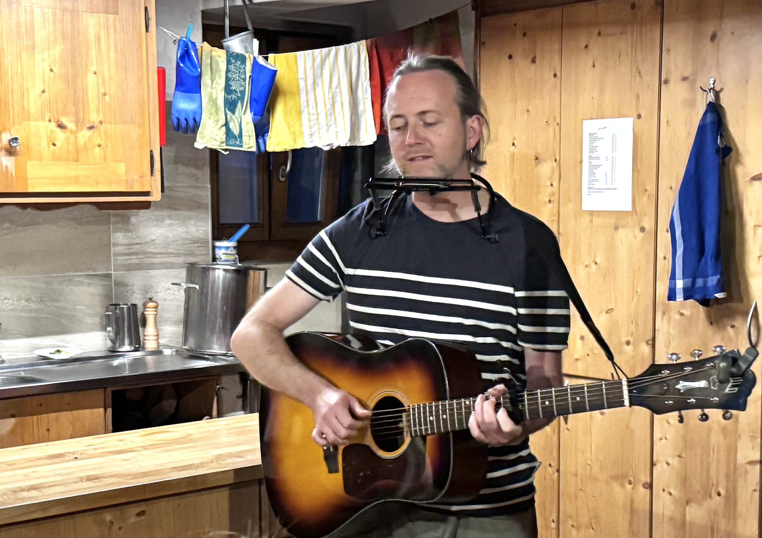 IMG_4326 Mann spielt Gitarre mit Mundharmonika in gemütlicher Holzküche, im Hintergrund bunte Handtücher und Küchenutensilien.